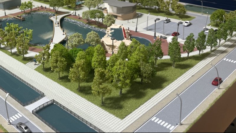 Architectural masterplan scale model: a tree-lined park with a lake and a footbridge, a car park beyond, and a canal and road along the near edge