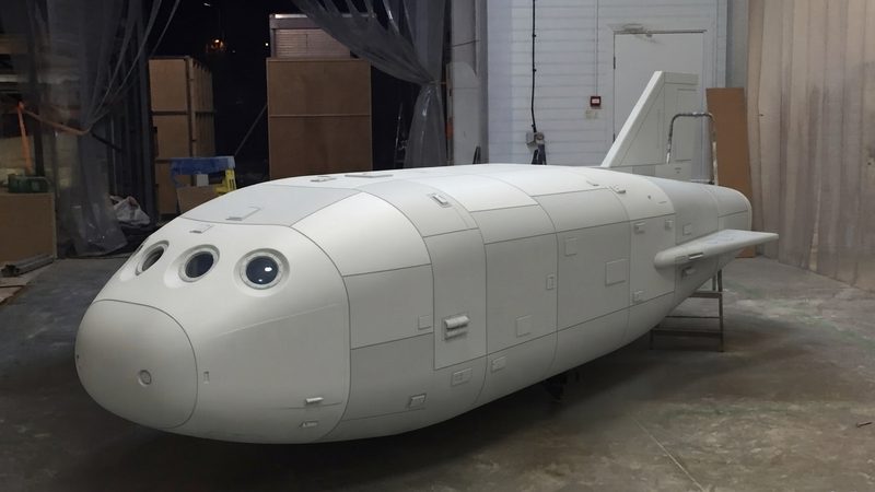 A large white panelled spacecraft prop standing in the middle of a workshop floor