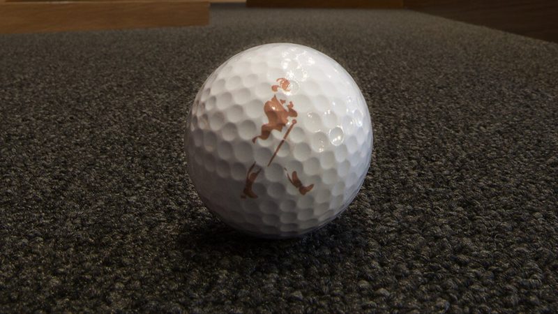A white golf ball marked with a copper logo on grey carpet at an exhibition