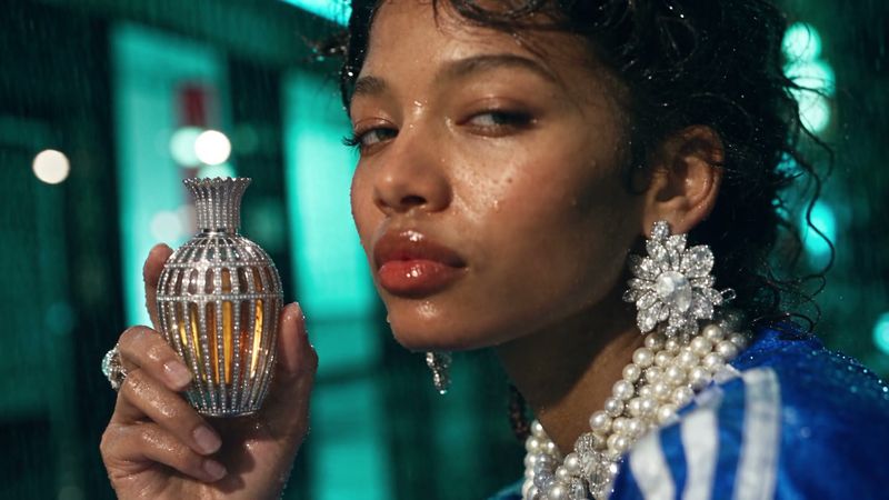 Close view of a woman's wet face and pearl necklace as she holds a crystal-set flacon up in the rain.