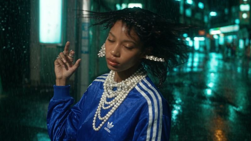 A woman in a blue tracksuit top and layered pearls tilts her head in the rain, her hair swinging, neon signs behind her.