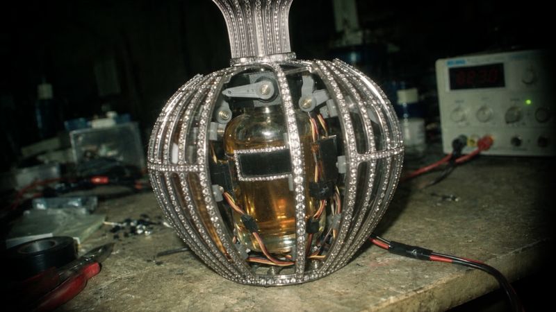A crystal-set flacon with its cage opened to show the servo mechanism and wiring inside, on a workbench beside a power supply.