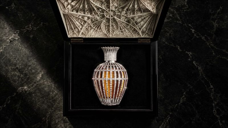 A crystal-set flacon standing in an open black presentation case with a carved white lid, on dark marble.